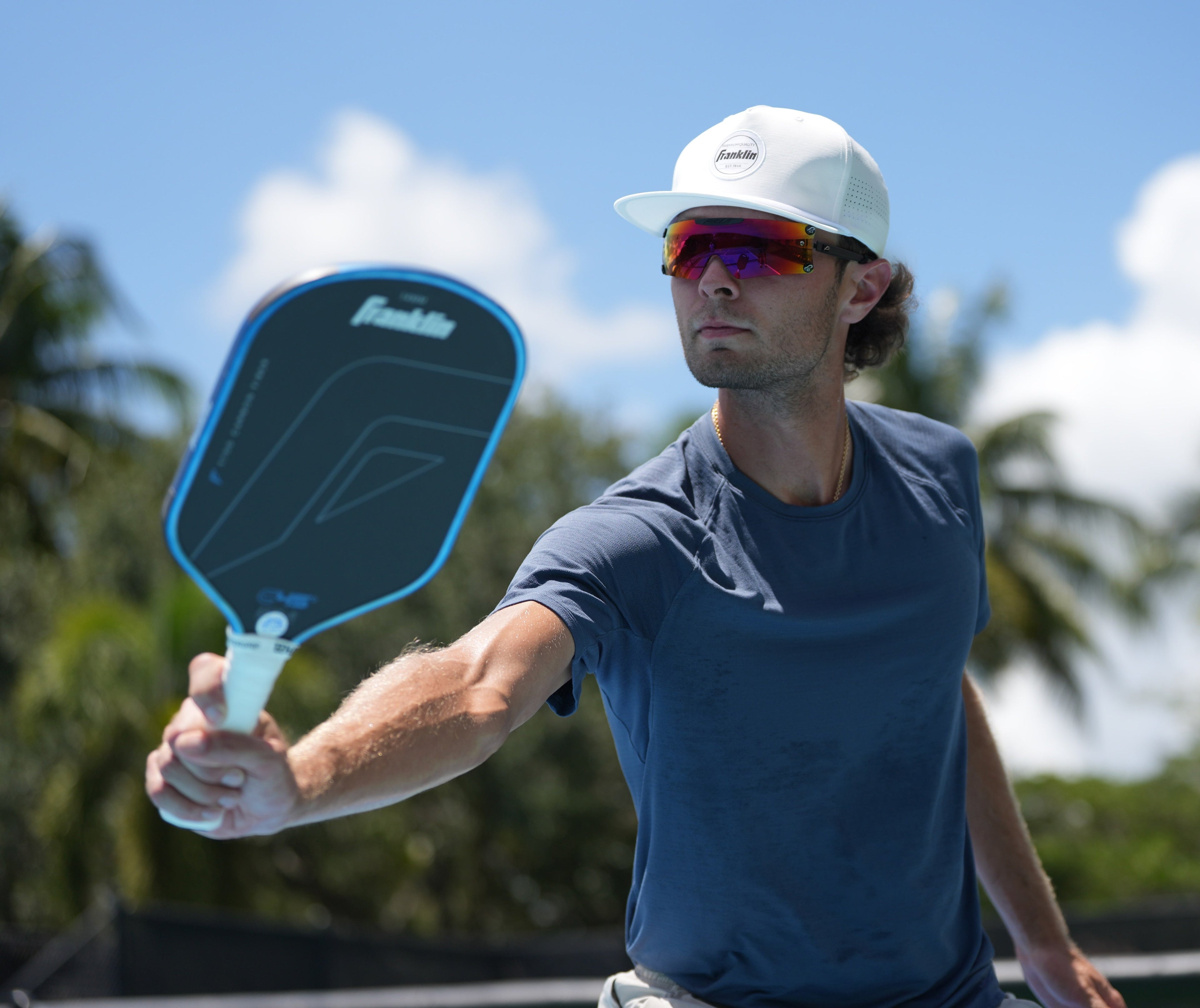 Man holding a pickleball paddle with a blue and black design, wearing a white cap and Pilla glasses, outdoors.